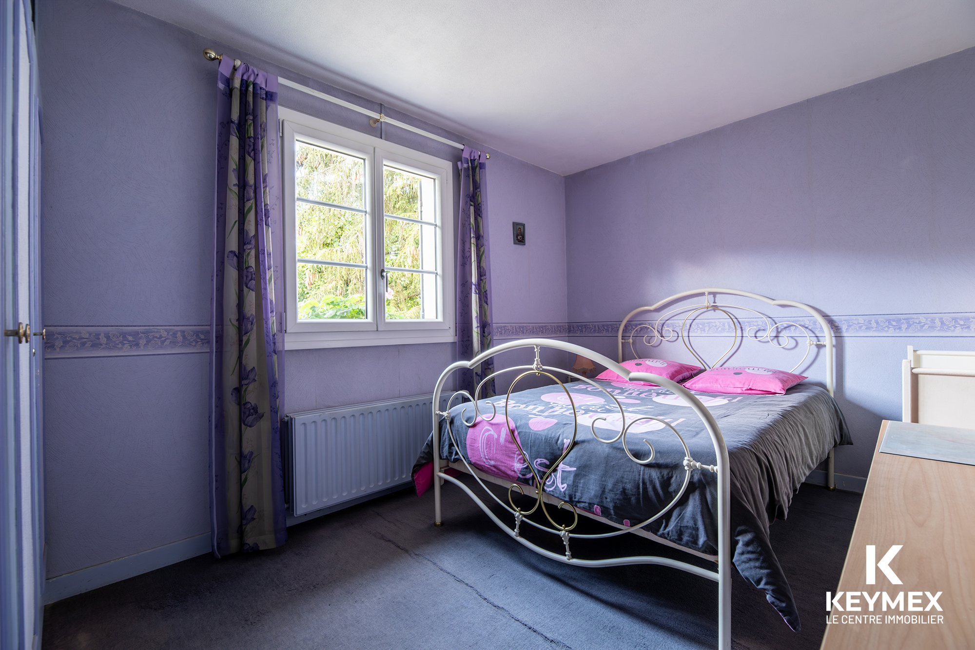 Chambre mauve avec lit double et fenêtre ensoleillée.