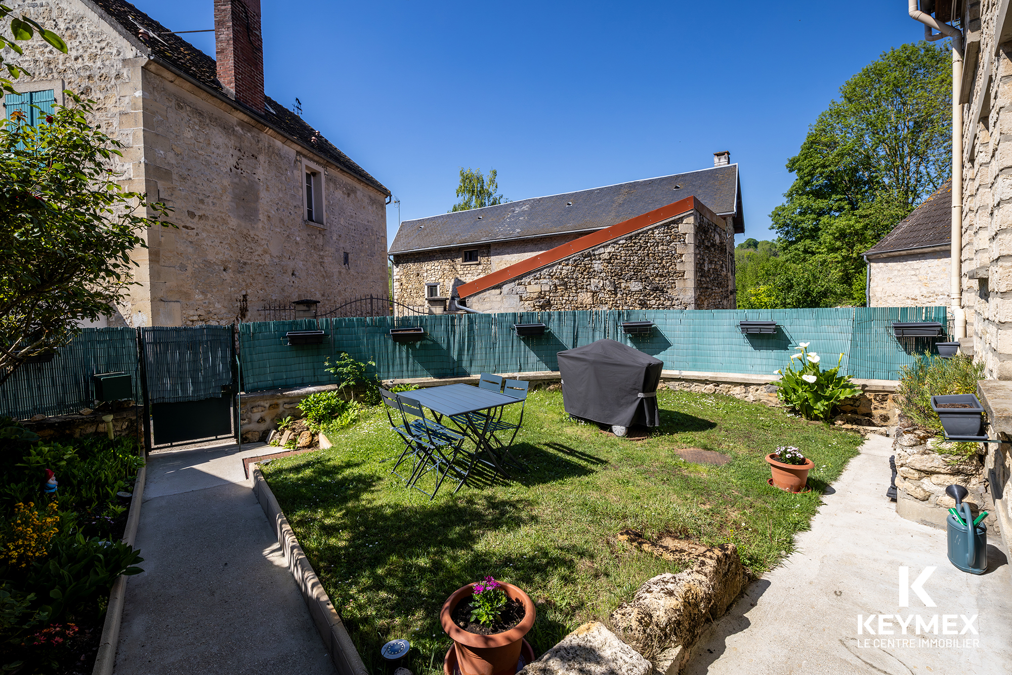 Jardin clôturé avec table et barbecue, maisons en pierre.