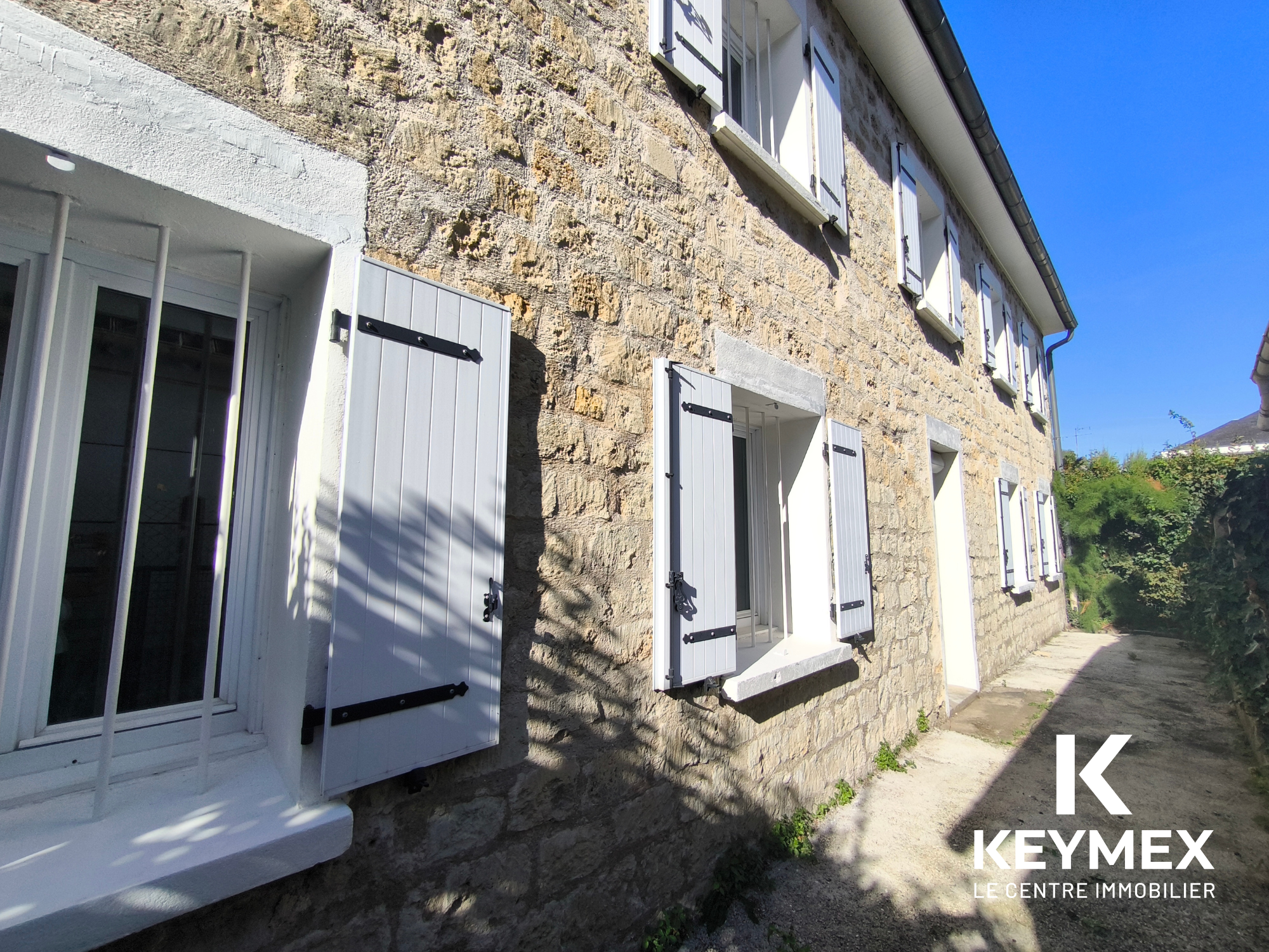 Maison en pierre avec volets blancs, façade ensoleillée.